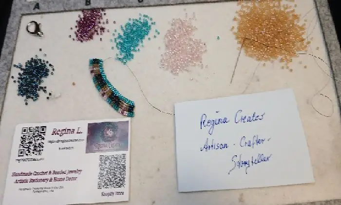 Regina Creates beading mat with several different colored delica beads, a project in progress, a business card, and a hand-written note.