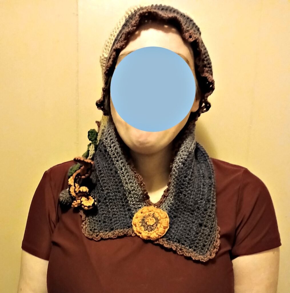 A crochet elf hat, perfect for fantasy wear, shown on a model with the face blocked out from the front view. It features a permanently closed attached neck warmer adorned with a crochet flower. Made in earthy tones using various yarn blends, with most of the material being acrylic yarn.