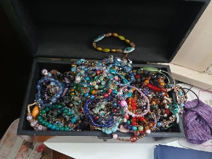 A top-down photograph looking into an open black jewelry box overflowing with a vibrant, dense collection of handmade beaded bracelets. The hoard includes dozens of bracelets featuring colorful glass beads, pearls, charms, metal spacers, and unique focal points, highlighting years of eclectic jewelry creation.