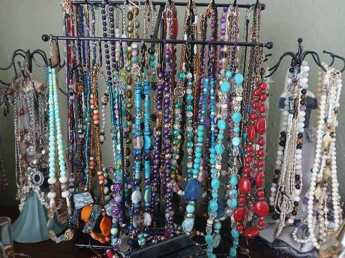 A close-up shot of several black metal jewelry display stands, densely packed and hanging with countless handmade beaded necklaces. The collection is highly varied, showcasing a multitude of colors, bead types, lengths, and styles, representing a large body of unique, artisan-crafted wearable art.