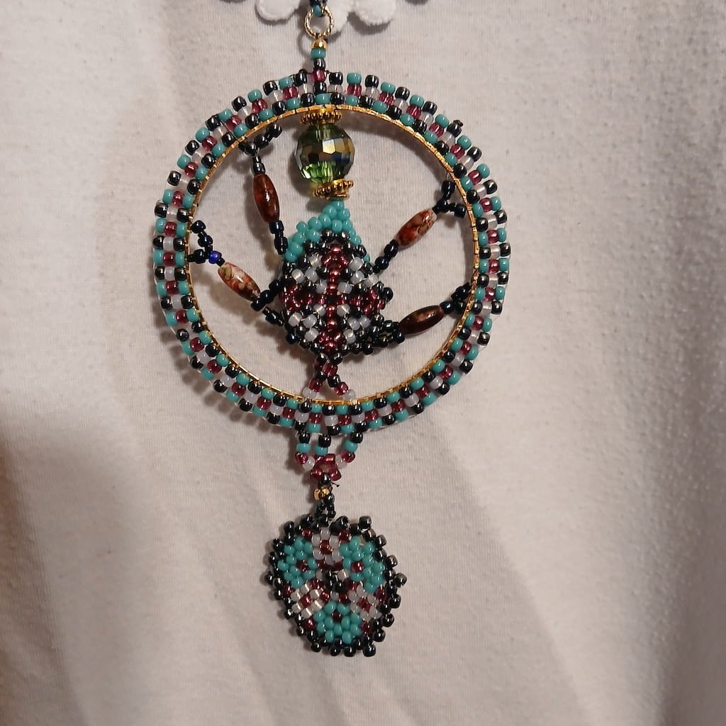 Handcrafted beaded Fantasy Fae Jewelry pendant necklace featuring an intricate circular design woven with teal, burgundy, black, and white seed beads on a gold frame, enclosing a stylized goddess figure with a green crystal head, and finished with a dangling beaded shield element, styled as a unique Renaissance Faire necklace accessory.