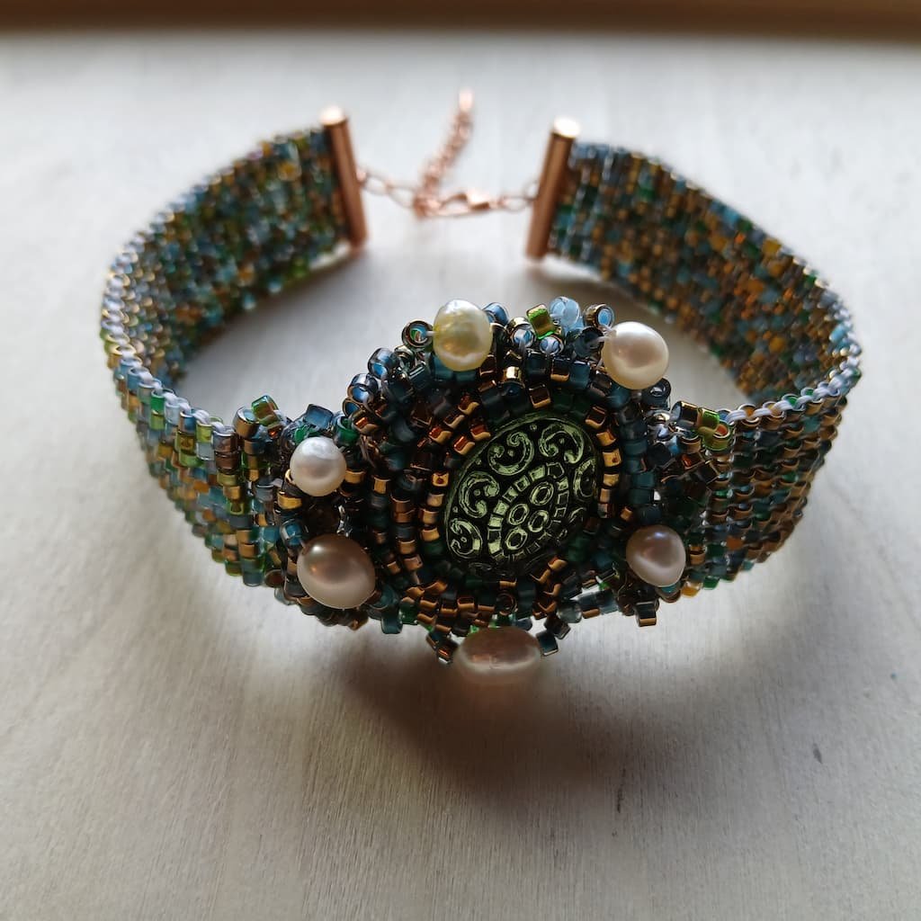 Handcrafted bead-woven Fantasy Fae Jewelry cuff bracelet featuring an intricate mix of metallic bronze, forest green, and teal seed beads, accented with luminous freshwater pearls surrounding a circular green cabochon centerpiece, designed as a nickel-free Renaissance Faire accessory.
