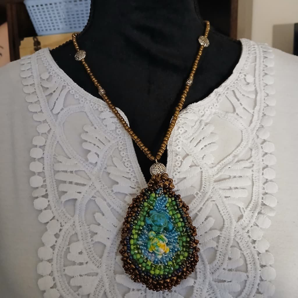 Handcrafted Sea and Earth Fantasy Fae Jewelry teardrop pendant necklace featuring a turtle motif intricately woven with shades of ocean blue, bright green, and yellow seed beads, framed by a thick border of metallic bronze beads and hung on a matching bronze beaded chain with silver filigree accents, perfect as a unique Renaissance Faire necklace.