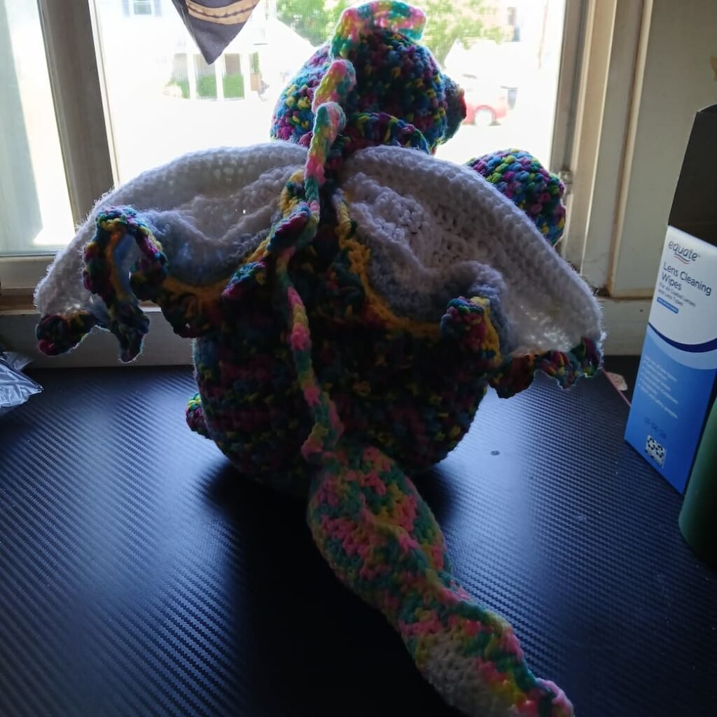 Rainbow crochet dragon toy with white inner wings sitting on a dark surface by a window.